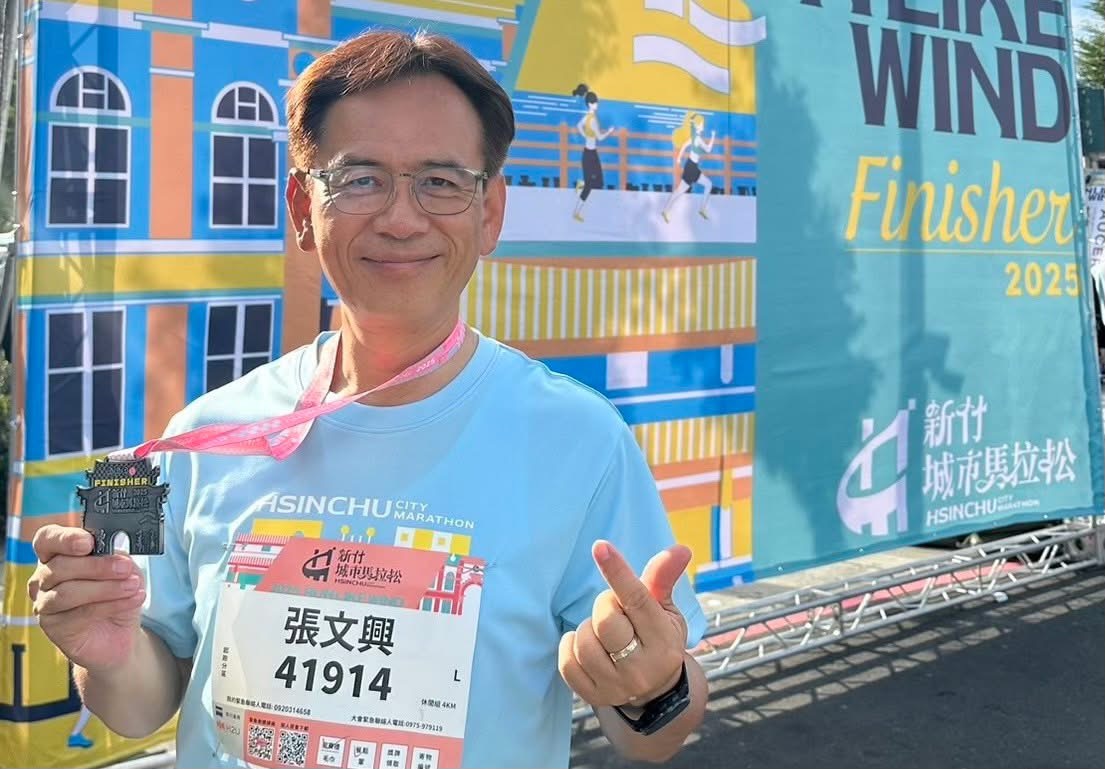 張文興理事長於 2025 新竹城市馬拉松順利完賽,並展示象徵賽事榮耀的迎曦門獎牌,背景為本屆大會主視覺,清晨陽光映照出路跑成就與城市活力。(圖/記者張家燁翻攝)