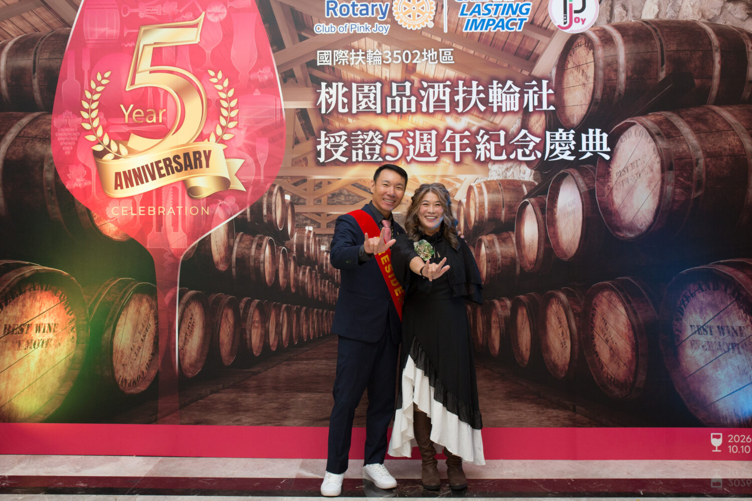 桃園品酒扶輪社社長蔡嘉益(Bruce)與夫人(Belle)於五週年授證慶典紀念背板前合影,留下溫馨時刻,象徵以酒會友、以心相聚的深厚扶輪情誼。(圖/記者 趙靜姸翻攝)