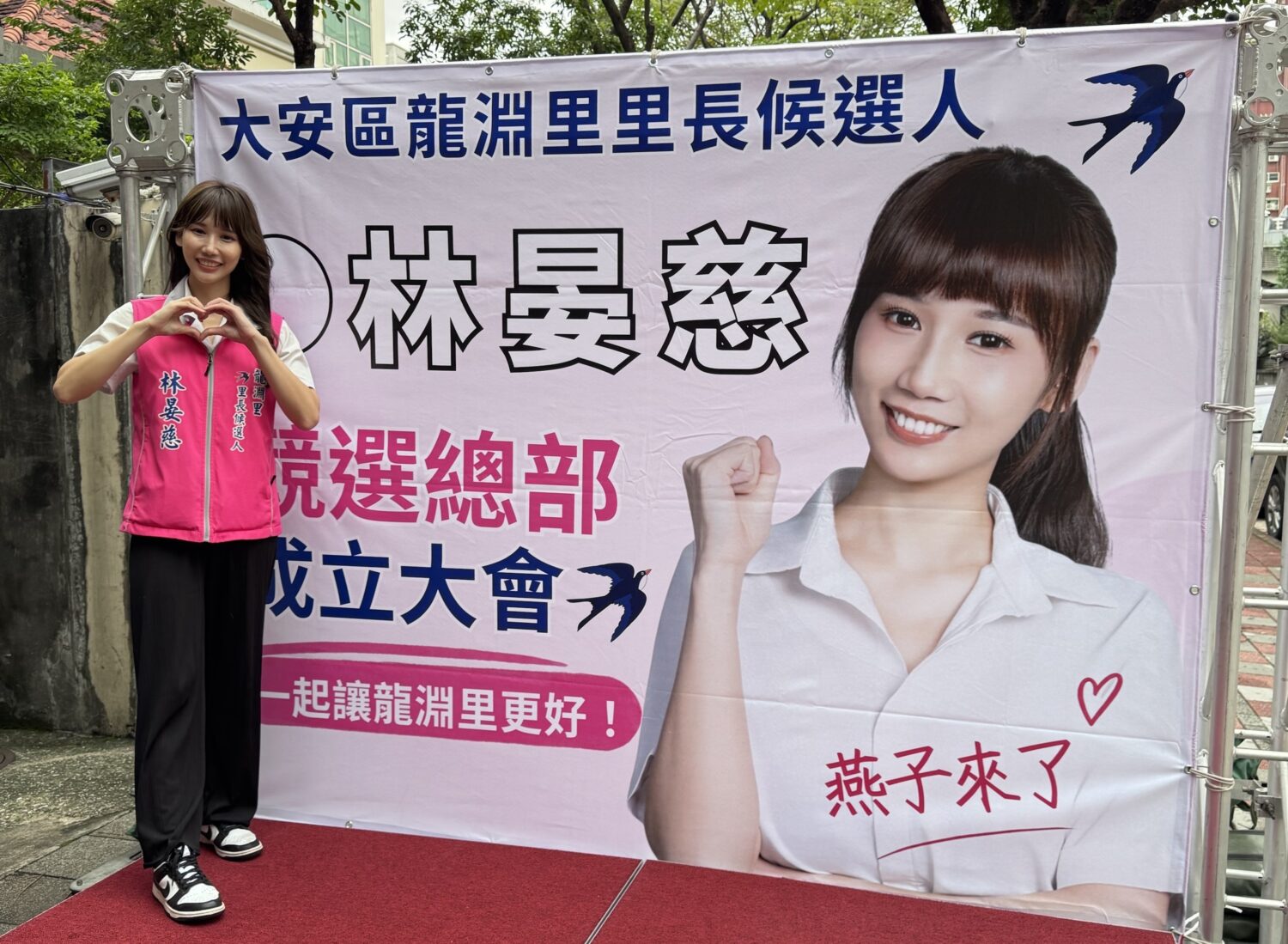 台北市大安區龍淵里里長候選人林晏慈，今（25）日舉辦競選總部成立大會，於競選主視覺布條前比出愛心手勢，展現親切形象。（圖／記者張家燁拍攝）